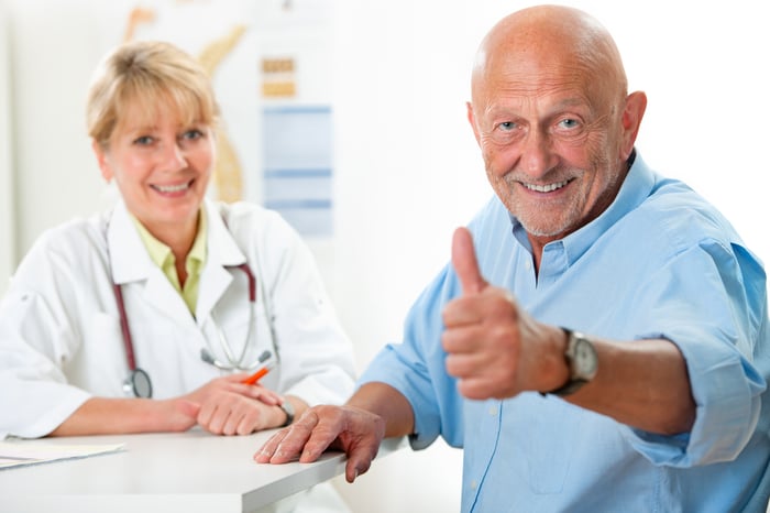 Older man sitting with health care professional, looking at viewer and smiling, giving thumbs-up.