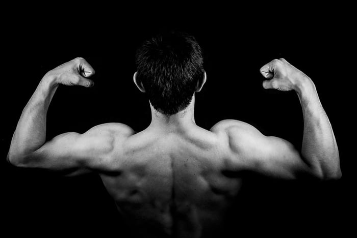 Black and white shot of the back of a man, making muscles, showing off biceps and back muscles