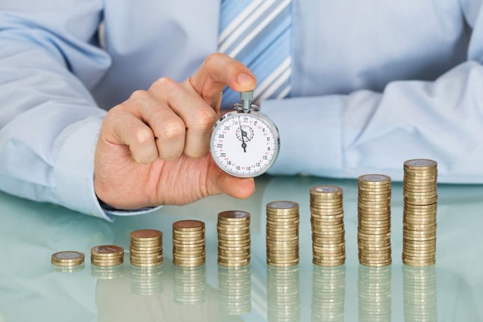 Businessman holding a stop watch in front of an ascending stack of coins, symbolizing long-term growth. 