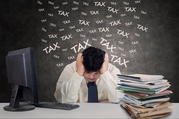 Man at desk with head in hands, near pile of folders, with the word "tax" written many times on a blackboard behind him.