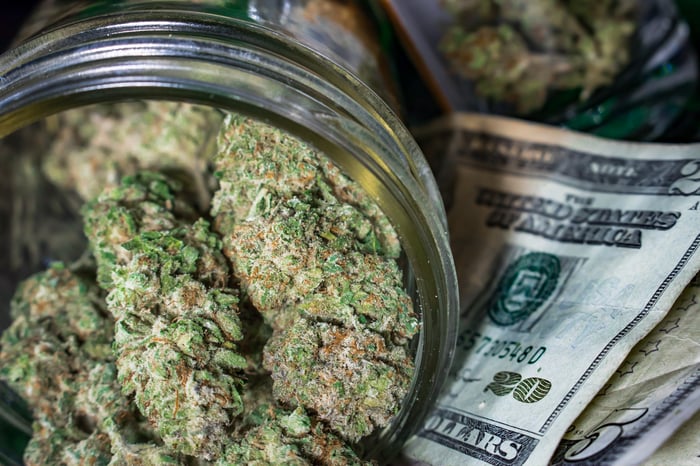 Marijuana buds in a jar sitting atop a pile of money. 