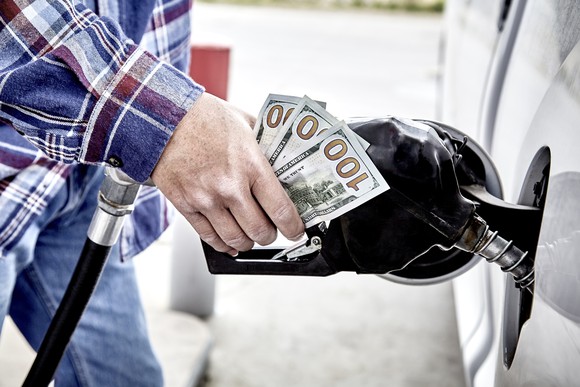 Man's hand holding three hundred US dollars and gas nozzle while pumping gas into parked vehicle.