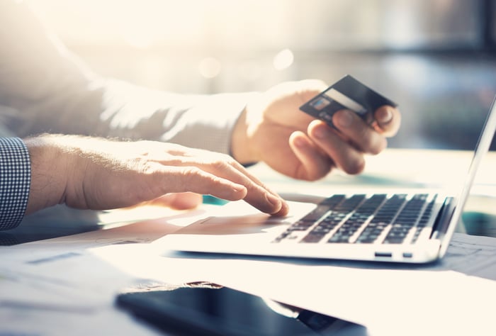 Hands in front of a laptop, one using the computer and another holding a credit card.