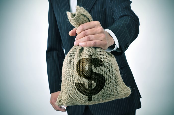 A man in a suit holds out a stuffed burlap bag with a dollar sign on it.