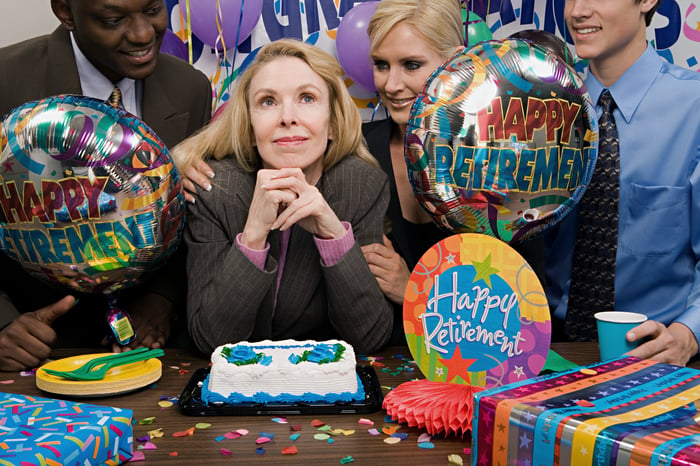 A woman smiles at her retirement party, with cake, balloons, and co-workers surrounding her.