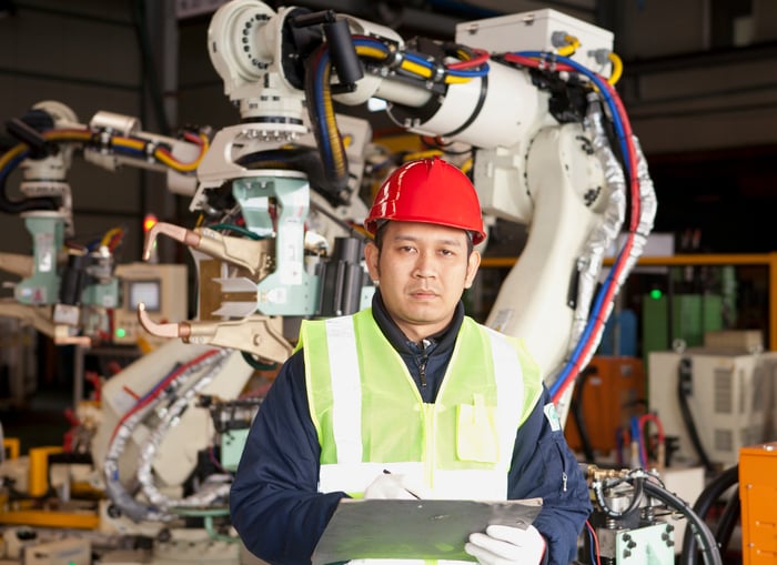 An engineer in a yellow vest and red hard hat holds a clipboard, with a robot in the background.