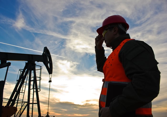 An oil pump jack next to an oil worker and a laptop.