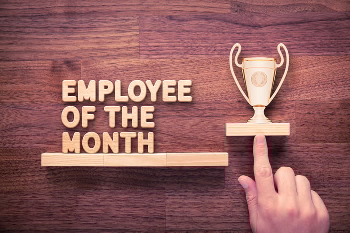 A finger points to a wooden trophy, which sits next to the words "Employee of the Month."