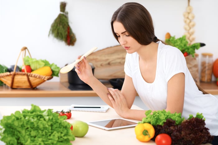 A woman, surrounded by fresh food in a kitchen and holding a wooden spoon in her hand, looks down at a tablet at the countertop.