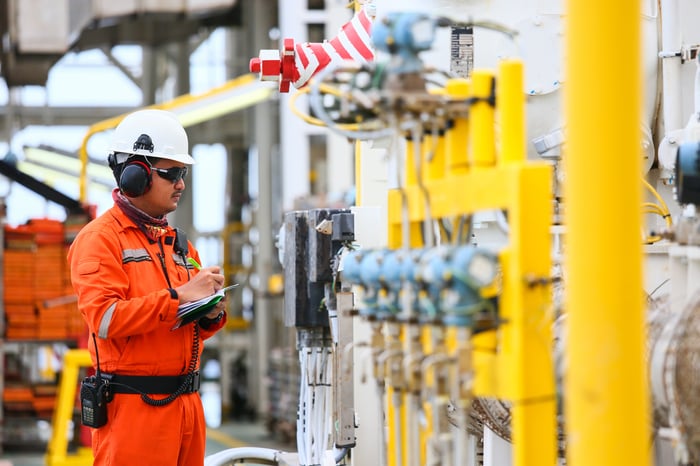 A man in an orange suit and a white hard hat holds a notebook and pen as he checks components at an oil and gas processing plant.