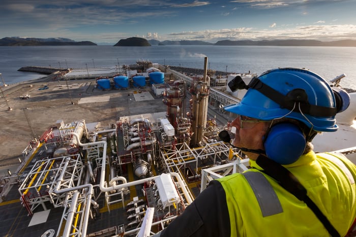 A man wearing blue ear protection, a blue hard hat, and a yellow vest, looking over oil and gas processing plant.