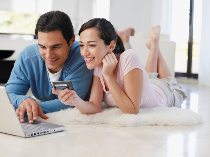 Man and woman lying on floor while types into laptop while woman holds credit card.