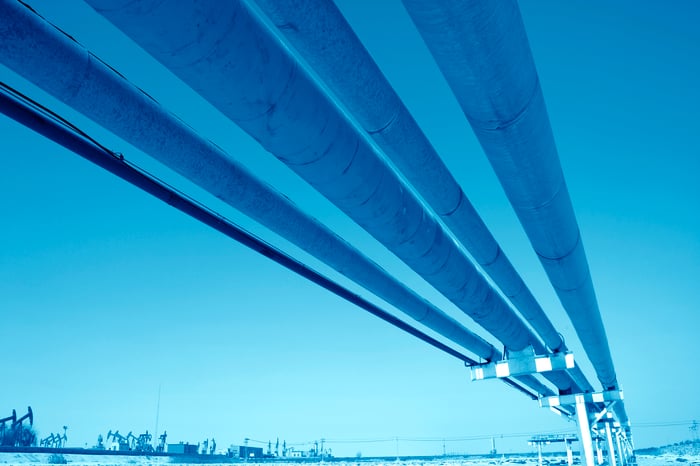 Several oil pipelines overhead with a blue background and oil pumpjacks in the foreground.