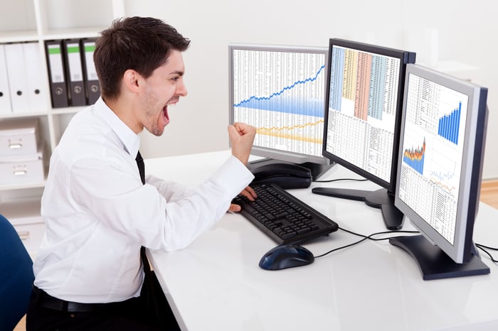 An investor cheering on big portfolio gains in front of three computer screens.