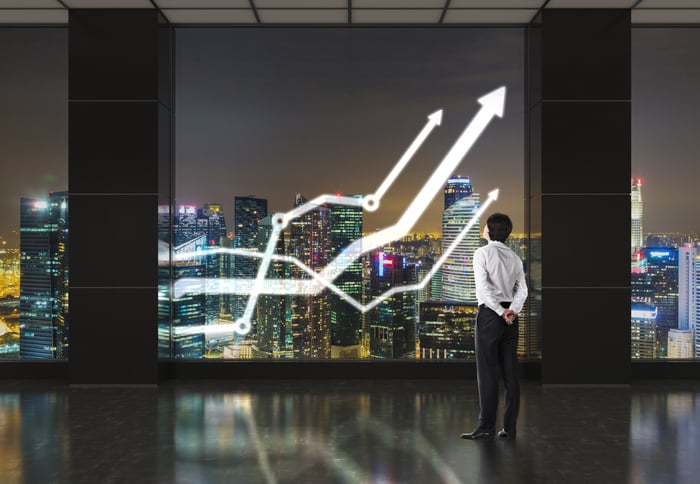 Well-dressed man looks at upward graph superimposed on window viewing cityscape at night.