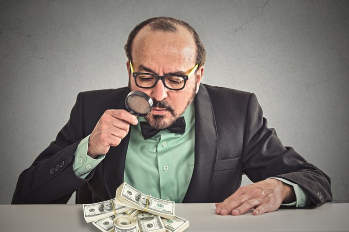 A man wearing a bowtie and suit jacket looking at pile of money with magnifying glass