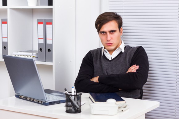 A displeased young worker with his arms folded at his desk.