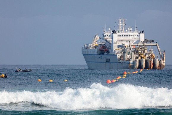 Orange buoys trail behind a ship laying the cable in the ocean. Some smaller single-person boats are in the water near the bigger ship.