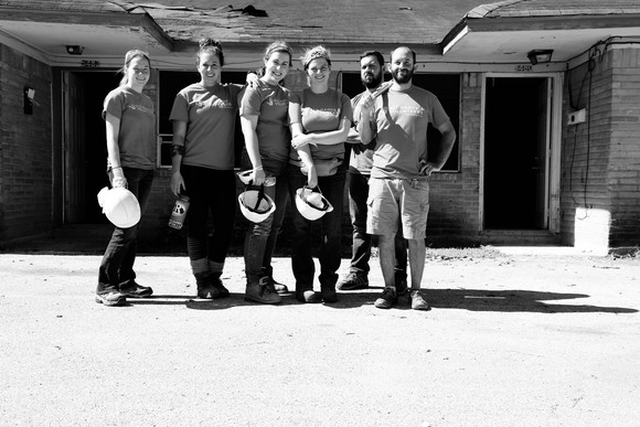 Six volunteers stand in front of a damaged house holding tools and construction helmets.