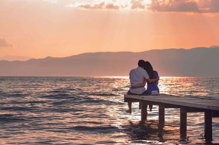 A couple sits on the end of a short dock on the water facing a sunset with their arms around each other. 