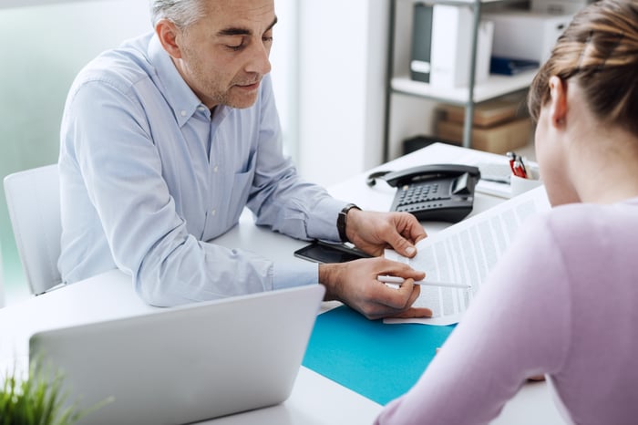 In an office setting, a man on one side of a desk reviews a document with a woman