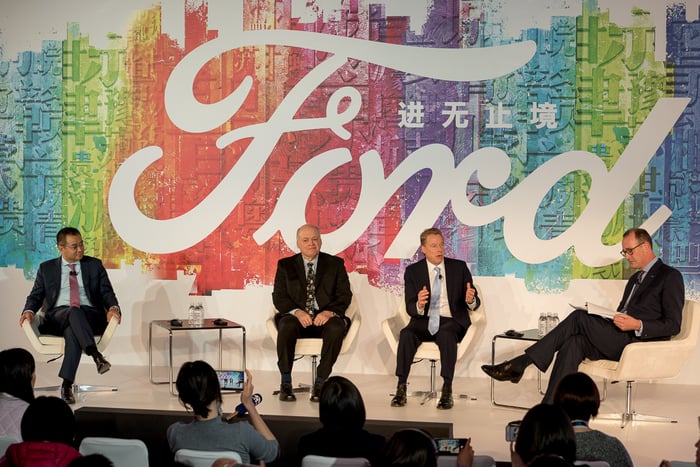 The four men are seated on a stage in front of a large rainbow-colored backdrop with a large Ford logo.