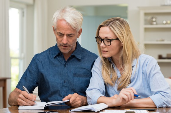 Older man writing in a notebook while woman looks on