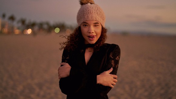 A model in a hat poses on beach crossing her arms and winking.