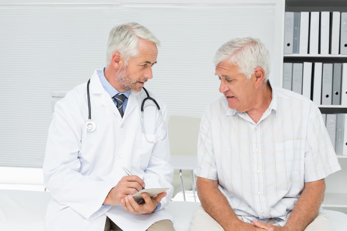 A senior male patient sits next to a doctor who holds a pen and paper in his hands.