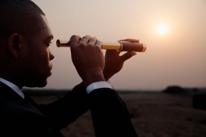 man in business suit looks through a telescope to a hazy horizon as the sun sets. 