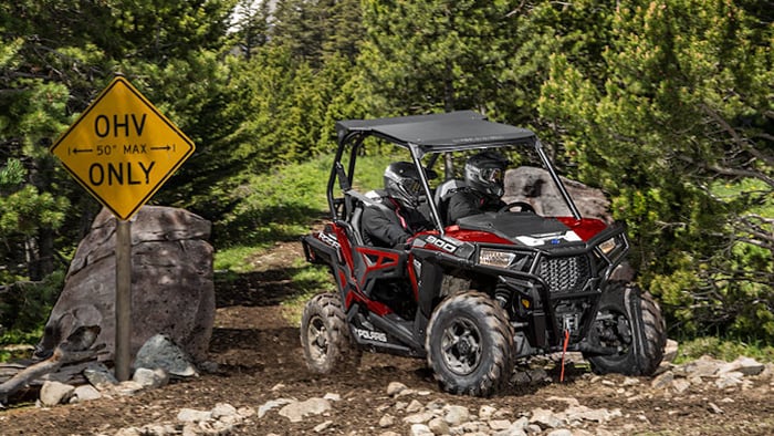 Two helmeted riders in a Polaris Industries RZR 900 on a wooded trail.