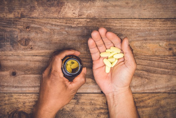 A pair of hands holding a bottle of vitamins and several vitamin tablets on a wooden table.