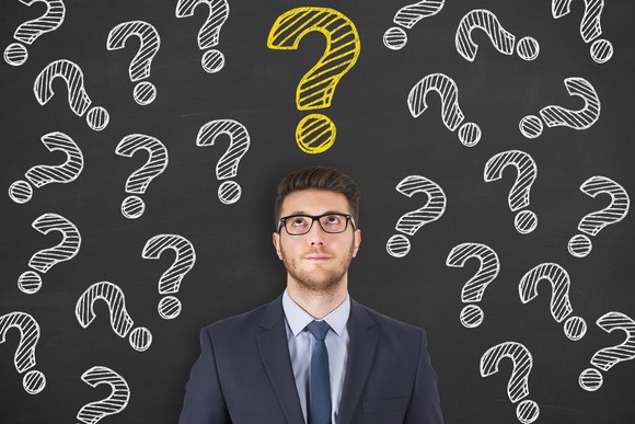 A man looks up at a blackboard decorated with question marks.