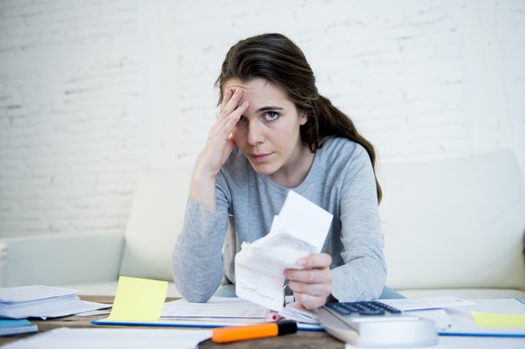 Woman sitting at a cluttered desk, putting a hand to her head while clutching a stack of papers.