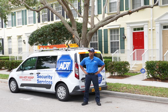 An ADT installer stands with his hands on his hips in front of an ADT van parked alongside a row of townhouses.