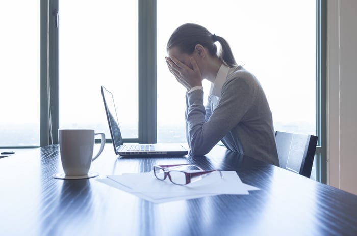 A professional woman sits in front of a laptop at a desk, with her face in her hands. 