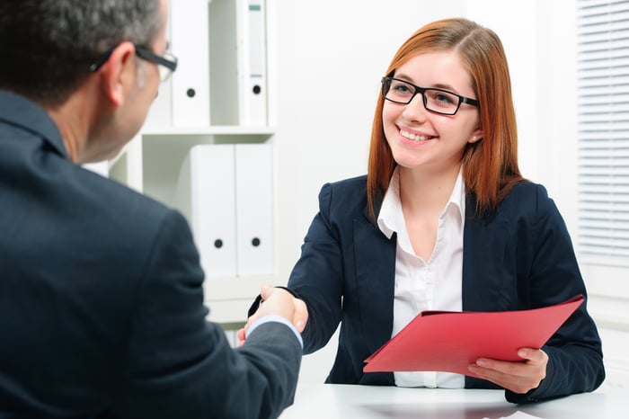 Professional young woman with a red folder in hand shaking hands with professional man in an office setting.