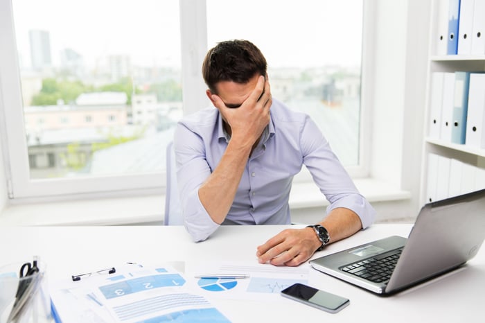 Man sitting at desk with laptop in front of him holding his face in the palm of his hand.