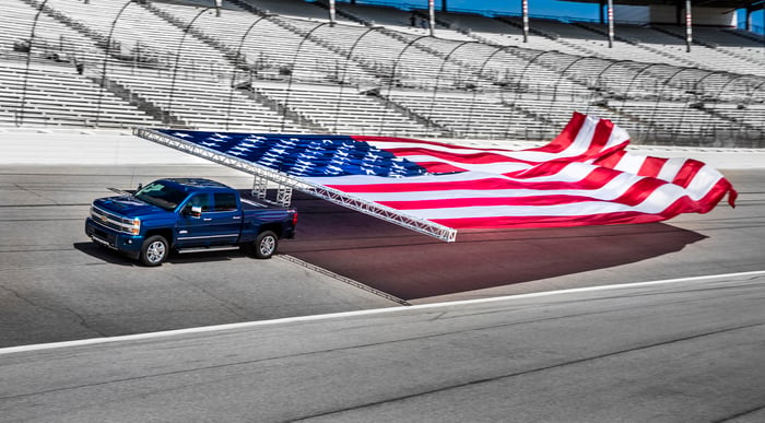 A blue Chevrolet Silverado truck hauling a large American flag on a racetrack.