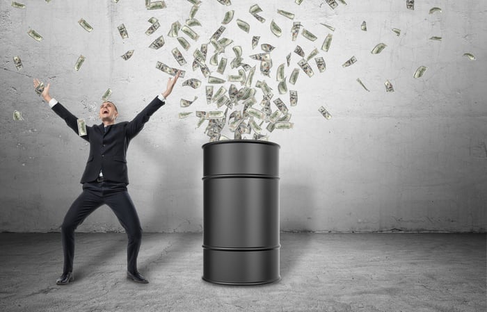 A black barrel on a gray floor with paper money flying out of it and a happy man in a black suit celebrating with outstretched arms next to it.