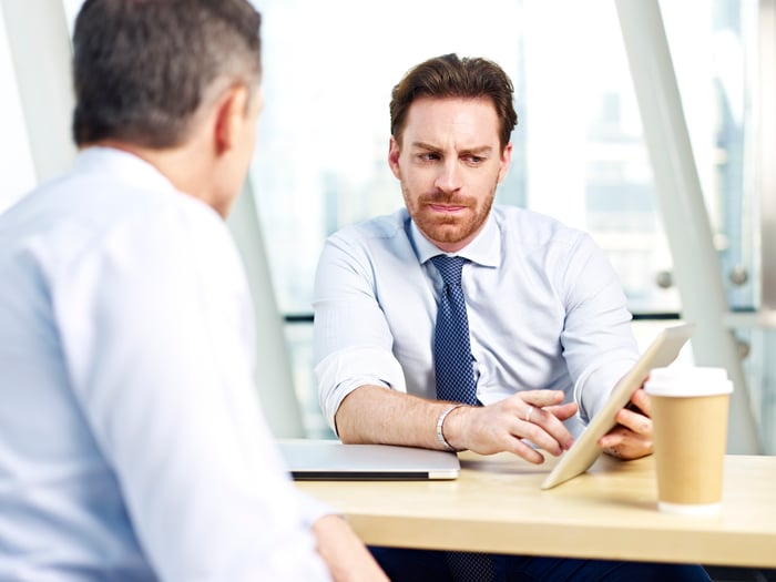 A man in a shirt and tie sitting across from another man, pointing to a tablet and frowning