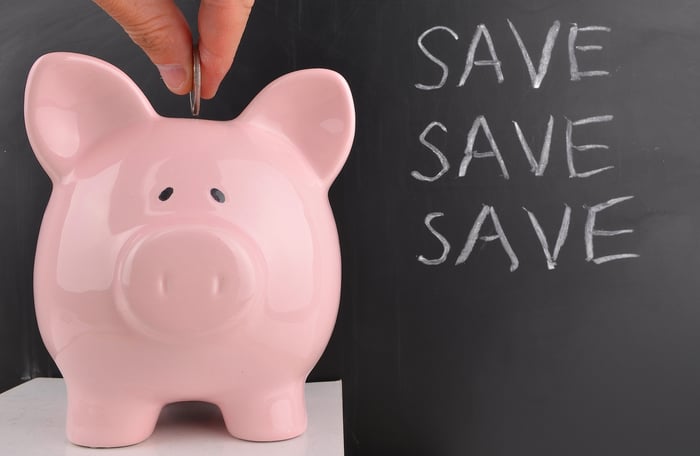 A coin being dropped into a pink piggy bank with the words save save save on a chalkboard behind it