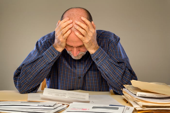 Senior man, with both hands holding his head, poring over stacks of papers, looking concerned.