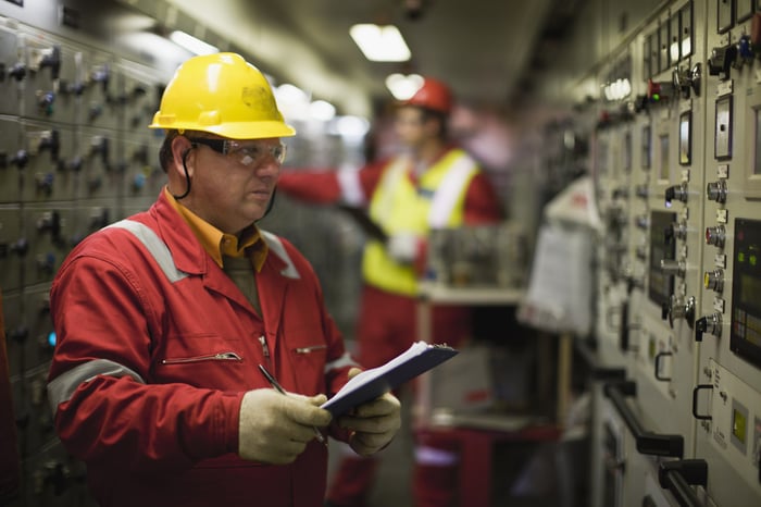 Man looking at industrial equipment while wearing a yellow hard hat and writing on a clipboard in his hands