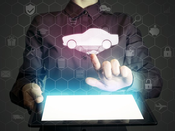 A man holding a tablet computer. Above the tablet, a simple image of a car floats in the air amid various icons related to money, shopping, and communications. He is clicking on the car image..