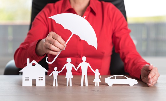 Woman in red shirt holding a paper umbrella over paper cutouts of a family, a house, and a car.