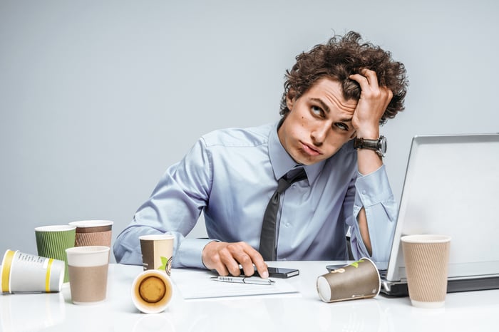 Stressed-looking man with his head propped up on his hand. There is a laptop and coffee cups on the table in front of him.