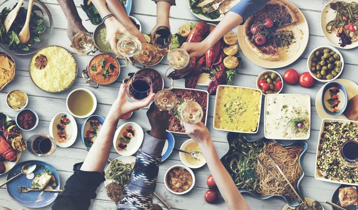 A table of various foods and wine. People off screen have their hands placed over the table, toasting.