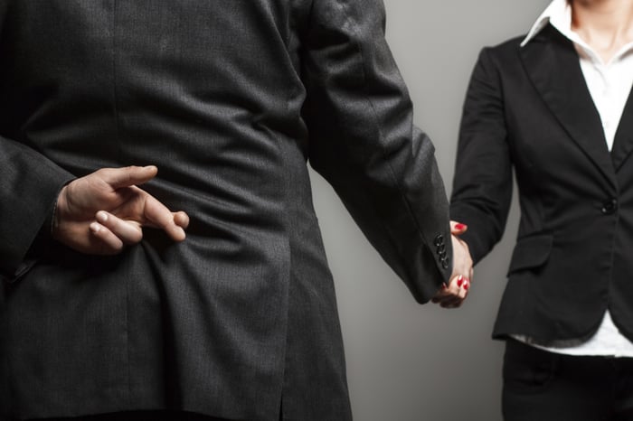 man in suit crossing fingers behind his back while shaking a womans hand