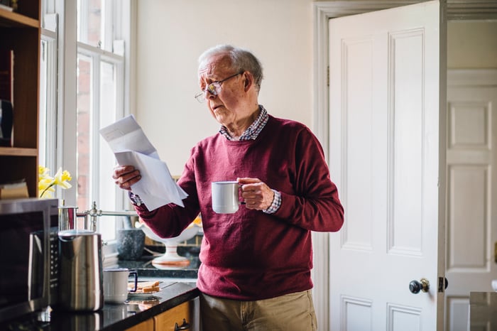 Senior man in a burgundy sweater holding a mug while reading paper bills